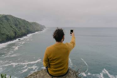 Rückansicht eines Mannes, der auf einem Felsen an der Küste sitzt und ein Smartphone-Foto auf der Insel Sao Miguel, Azoren, Portugal, macht - AFVF06306