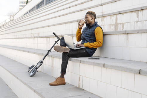 Young man with laptop and scooter sitting on stairs drinking coffee - JCZF00082