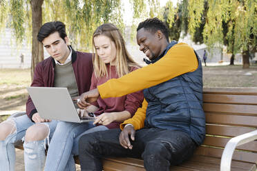 Freunde sitzen auf einer Parkbank und teilen sich einen Laptop - JCZF00096