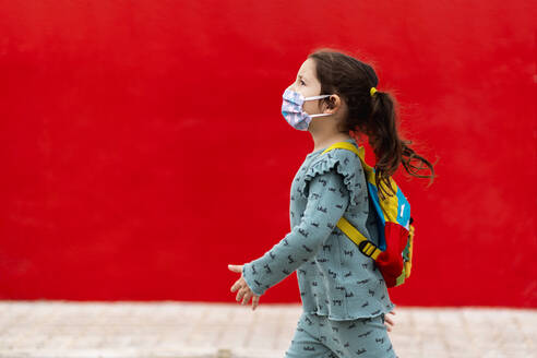Girl walking with backpack and mask along red wall - EGAF00112
