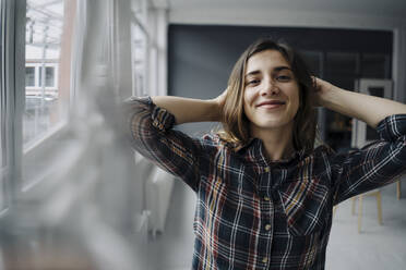 Porträt einer glücklichen jungen Frau mit den Händen hinter dem Kopf in einem Loft - JOSEF00790