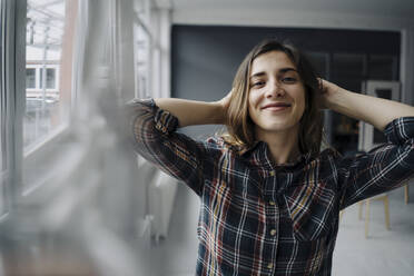 Portrait of happy young woman with hands behind head in a loft - JOSEF00790