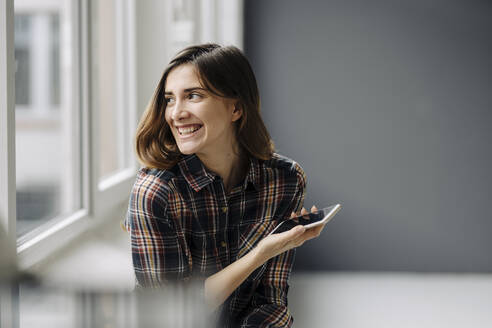 Portrait of laughing young woman with smartphone looking out of window - JOSEF00793