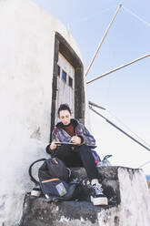 Young woman sitting at a building with book and pencil, Corvo, Azores, Portugal - FVSF00291