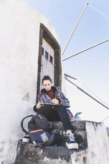 Junge Frau sitzt an einem Gebäude mit Buch und Bleistift, Corvo, Azoren, Portugal - FVSF00291