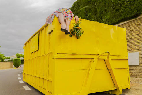 Woman lying on top of yellow container, holding buch of flowers - ERRF03854