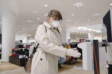 Woman with face mask and disposable gloves disinfecting her hands before shopping in a fashion store - AHSF02587