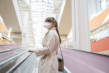 Woman with face mask and disposable gloves on an escalator in a shopping center - AHSF02593