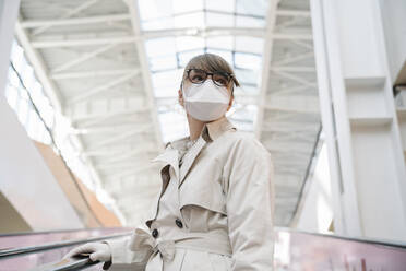 Frau mit Gesichtsmaske und Einweghandschuhen auf einer Rolltreppe in einem Einkaufszentrum - AHSF02594