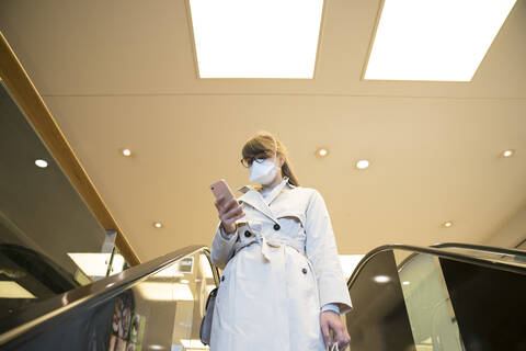 Woman with face mask using smartphone on an escalator in a shopping center stock photo