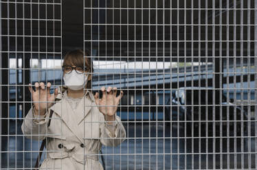 Woman wearing face mask standing behind grating in a car park - AHSF02610