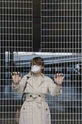 Woman wearing face mask standing behind grating in a car park - AHSF02611