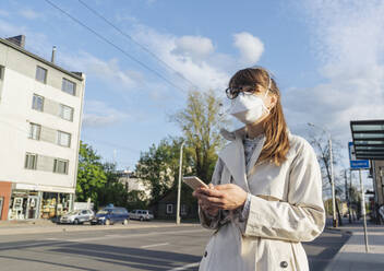 Frau mit Gesichtsmaske und Smartphone in der Hand wartet auf einen öffentlichen Verkehrsmittel in der Stadt - AHSF02617