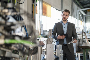 Junger Geschäftsmann mit Tablet und Werkstück an einer Maschine in einer Fabrik - DIGF11055