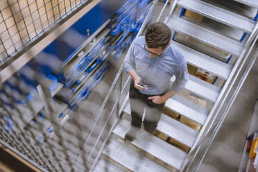 Businessman using smartphone walking down stairs in a factory - DIGF11150