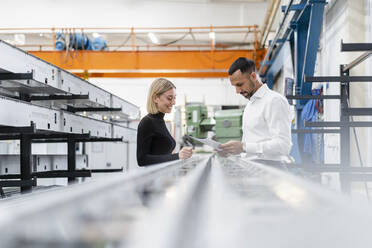 Businessman and woman with papers at metal rods in factory hall - DIGF11162