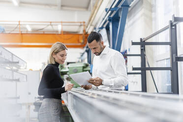 Geschäftsmann und Frau mit Papieren untersuchen Metallstangen in einer Fabrikhalle - DIGF11163
