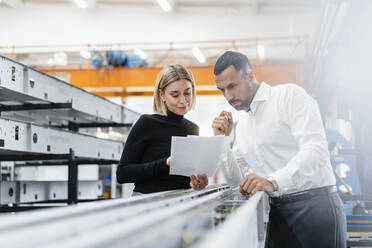 Geschäftsmann und Frau mit Papieren untersuchen Metallstangen in einer Fabrikhalle - DIGF11165