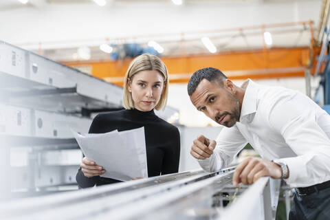 Geschäftsmann und Frau mit Papieren untersuchen Metallstangen in einer Fabrikhalle, lizenzfreies Stockfoto