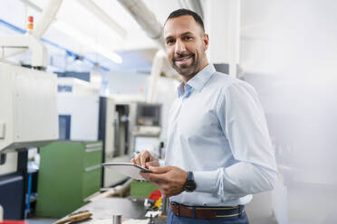 Portrait of smiling businessman holding tablet in factory hall - DIGF11185