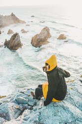 Junge Frau in warmer Kleidung sitzt auf einem Felsen am Strand von Ursa, Region Lisboa, Portugal - FVSF00318