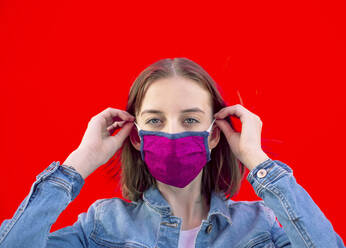 Portrait of teenage girl putting on protective mask against red background - BFRF02247