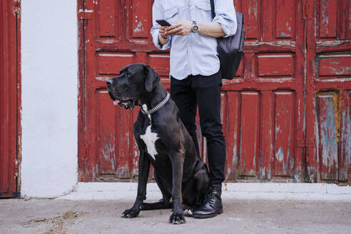 Crop-Ansicht eines jungen Mannes mit Handy, der mit seinem Hund vor einer alten roten Holztür wartet - EBBF00105