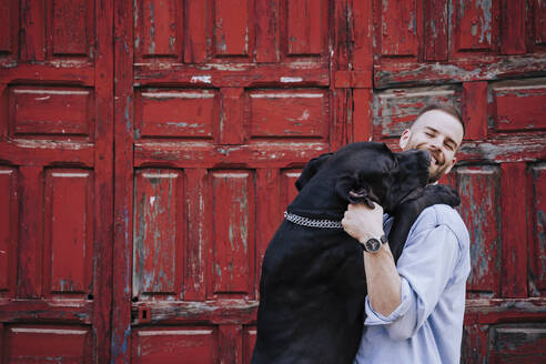 Porträt eines lachenden jungen Mannes und seines Hundes vor einer alten roten Holztür - EBBF00107