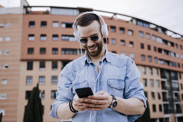 Porträt eines lächelnden jungen Mannes mit Kopfhörern, der auf sein Mobiltelefon schaut - EBBF00114