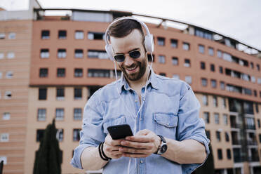 Portrait of smiling young man with headphones looking at cell phone - EBBF00114