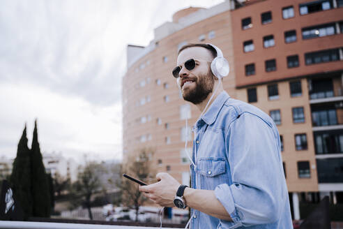 Porträt eines lächelnden jungen Mannes, der mit Kopfhörern und Smartphone im Freien Musik hört - EBBF00116