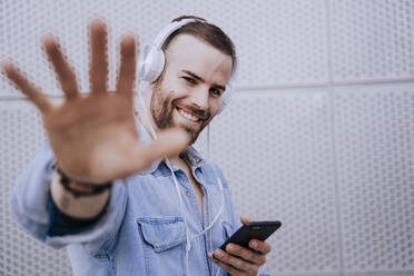 Porträt eines lächelnden jungen Mannes mit Kopfhörern und Smartphone, der die Hand hebt - EBBF00121