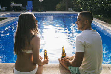 Glückliches junges Paar, das sich am Pool sitzend mit Rübenflaschen unterhält - ABZF03125
