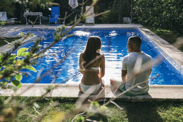 Rear view of young couple talking while sitting at poolside - ABZF03127