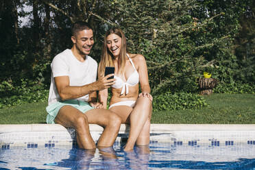 Smiling man showing smart phone to girlfriend while sitting against trees at poolside - ABZF03129