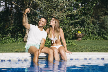 Smiling man taking selfie with girlfriend over smart phone while sitting against trees at poolside - ABZF03130