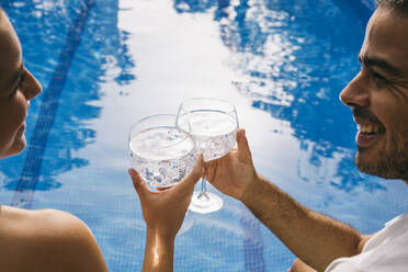 Fröhliches junges Paar, das sich beim Anstoßen mit Getränken am Swimmingpool ansieht - ABZF03132