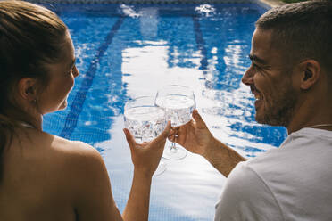 Cheerful young couple looking at each other while toasting glasses against swimming pool - ABZF03133