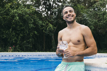 Lächelnder, hemdsärmeliger junger Mann, der ein Getränk in der Hand hält und wegschaut, während er im Schwimmbad vor Bäumen steht - ABZF03134