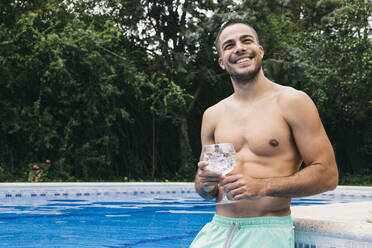 Smiling shirtless young man holding drink looking away while standing against trees in swimming pool - ABZF03134