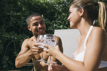 Smiling young man toasting drink with girlfriend while sitting on deck chair - ABZF03140