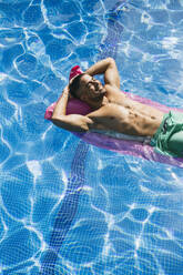 Hemdenloser hübscher junger Mann mit Händen hinter dem Kopf entspannt auf einer Luftmatratze im Schwimmbad - ABZF03144
