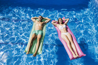 Lächelndes junges Paar mit Händen hinter dem Kopf, das sich auf Luftmatratzen im Schwimmbad entspannt - ABZF03146