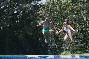 Cheerful young couple holding hands and jumping in swimming pool - ABZF03147