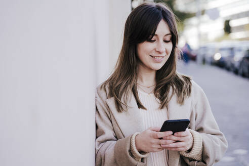 Porträt einer lächelnden jungen Frau, die an der Wand lehnt und auf ihr Smartphone schaut - EBBF00139