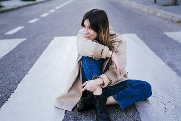 Portrait of smiling young woman sitting on zebra crossing - EBBF00156