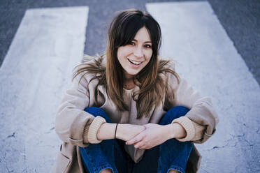 Portrait of smiling young woman sitting on zebra crossing - EBBF00157