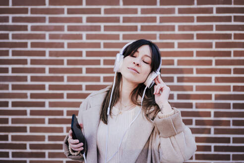Porträt einer Frau mit geschlossenen Augen, die mit Kopfhörern und Smartphone vor einer Backsteinmauer Musik hört - EBBF00164