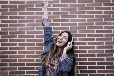 Portrait of happy woman with eyes closed listening music with headphones in front of brick wall - EBBF00169