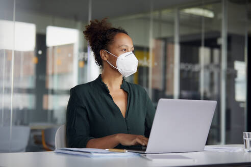 Portrait of businesswoman wearing protective mask working on laptop at counter looking at distance - RBF07708